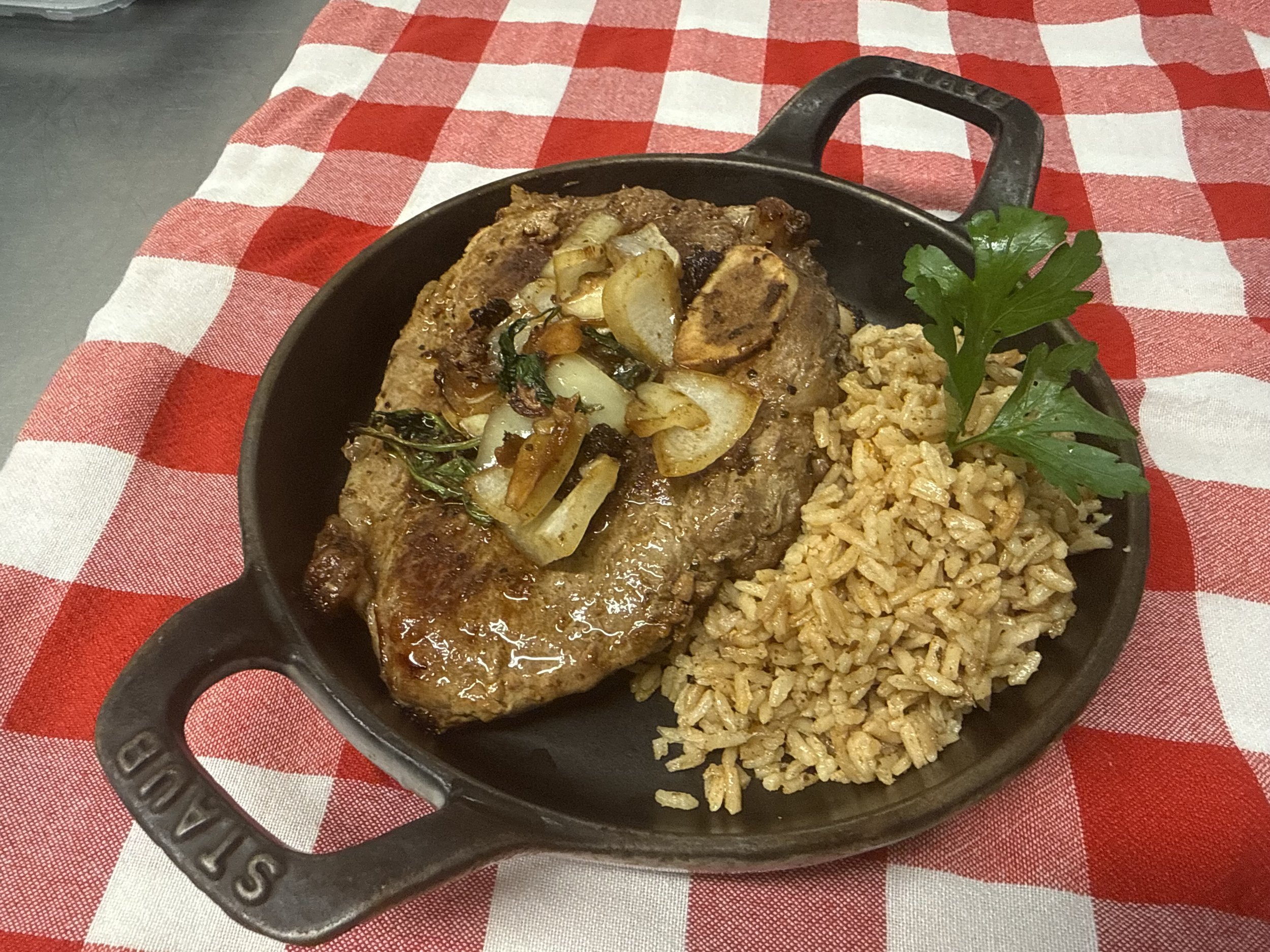 Juicy grilled steak topped with sautéed onions and herbs, served alongside flavorful seasoned rice, presented in a cast iron skillet on a red and white checkered tablecloth.