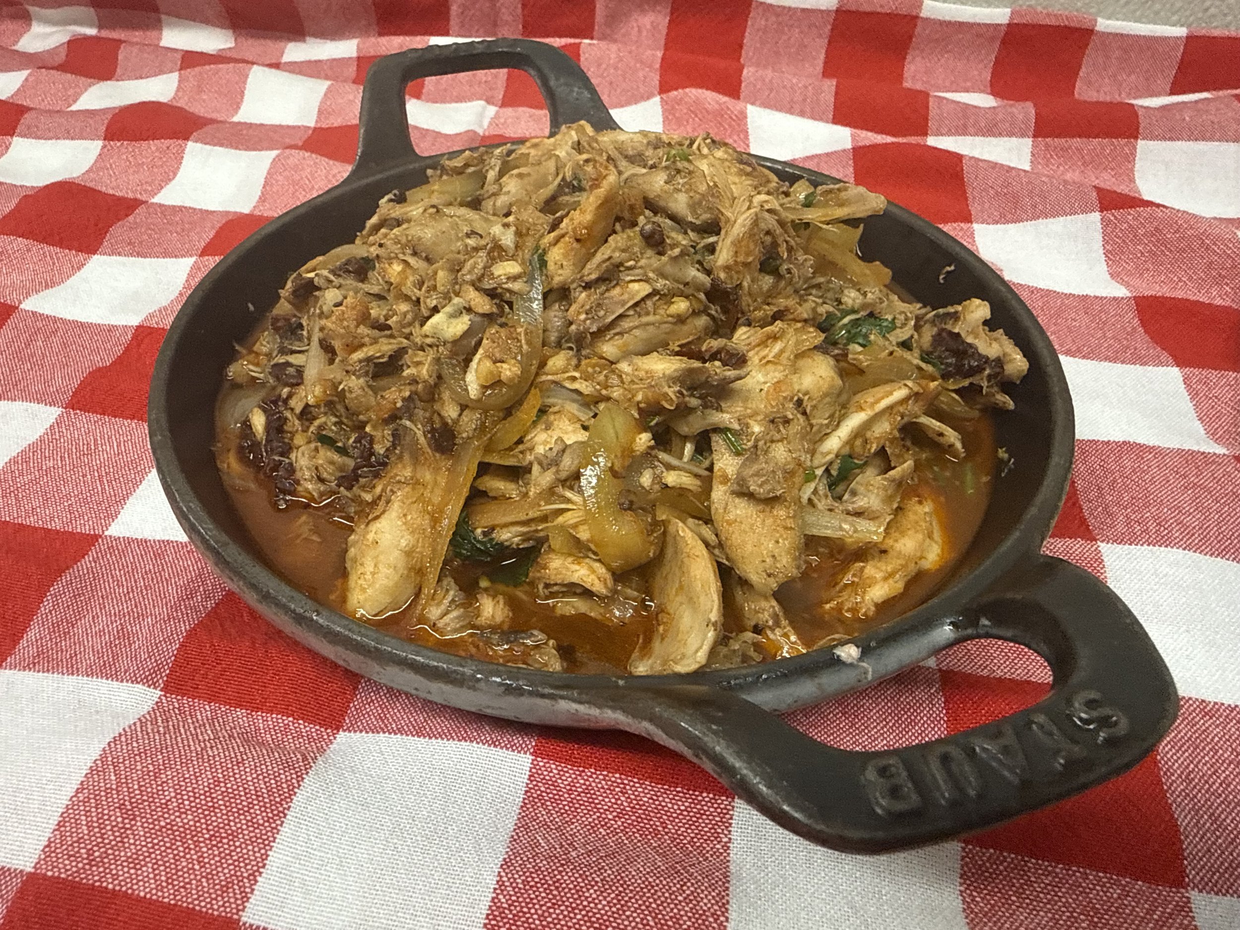Cast iron skillet filled with shredded chicken, onions, and spices, served on a red and white checkered tablecloth, highlighting a delicious homemade dish.