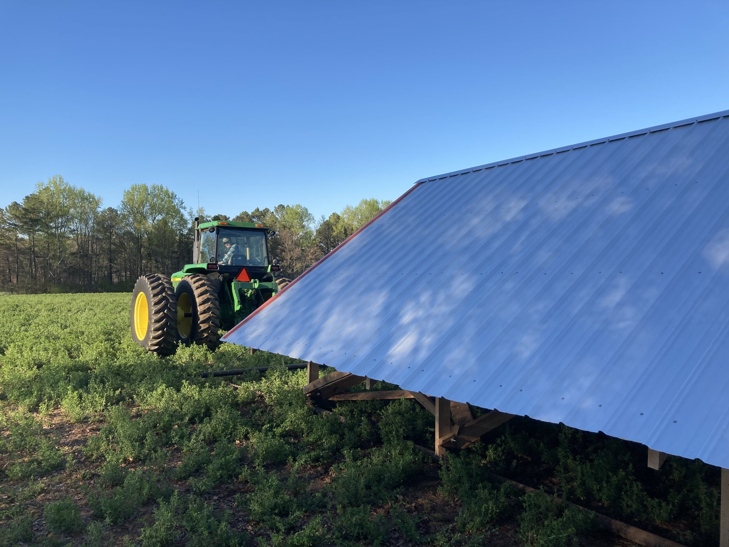 Green tractor working in a lush field under a clear blue sky, with a metal shed in the foreground, showcasing agricultural activity and equipment in a rural setting.