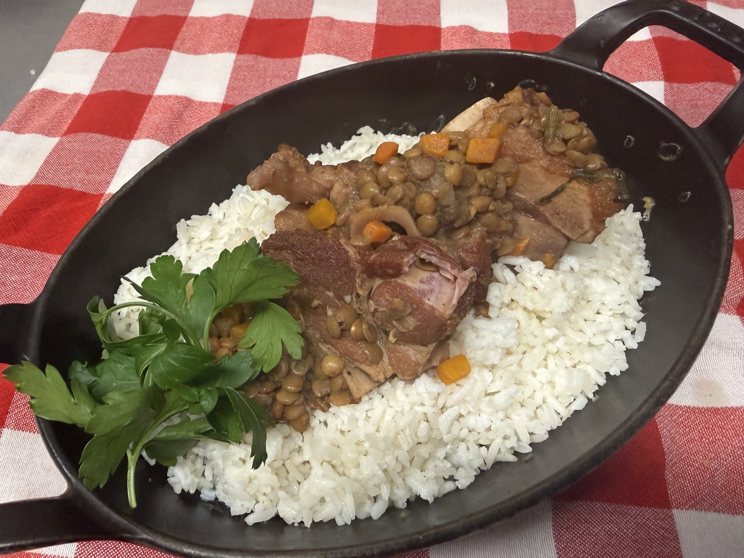 A cast iron skillet filled with tender meat, lentils, and colorful vegetables served over fluffy white rice, garnished with fresh parsley, on a red and white checkered tablecloth.