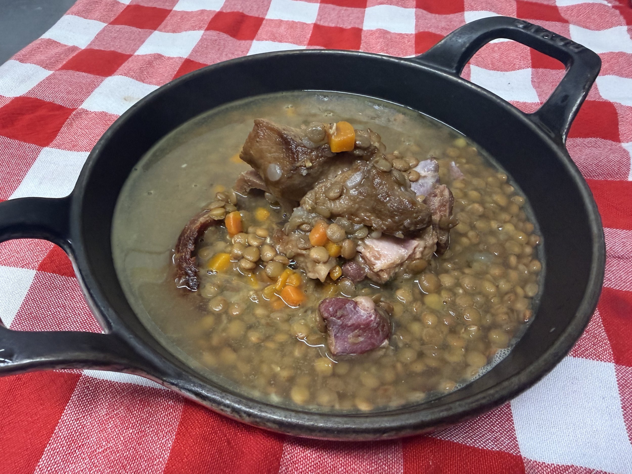 Bowl of lentil stew featuring tender pieces of meat and diced vegetables, served in a black cast iron dish on a red and white checkered tablecloth.